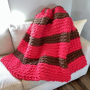 May include: A chunky knit blanket in a vibrant pink with wide brown stripes. The blanket is draped over a light gray sofa, showcasing its soft texture and bold color contrast. This cozy throw blanket adds a pop of color to any living space.
