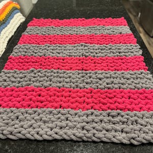 May include: A handmade, chunky knit blanket with alternating stripes of gray and bright pink. The thick yarn creates a textured surface, perfect for a cozy home decor item. The blanket is displayed on a dark countertop.