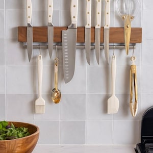 May include: A kitchen knife set with white handles and stainless steel blades, displayed on a wooden magnetic strip. Includes a chef's knife, paring knives, and a pair of gold-toned scissors. Also shown are a silicone spatula, a brush, and measuring spoons.