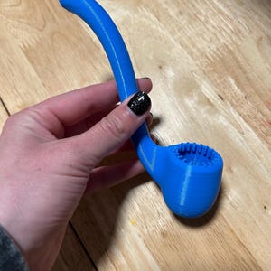 Puede incluir: Un objeto azul brillante en forma de pipa, con un tallo curvo y un cuenco texturizado. El objeto, probablemente impreso en 3D, se sostiene en una mano. El interior del cuenco tiene un diseño estriado. La forma de la pipa recuerda a una pipa tradicional.