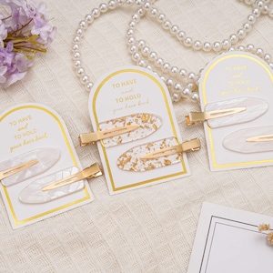 May include: Three sets of hair clips on display. Each set includes two oval-shaped clips with gold-coloured metal accents. The clips are white with gold leaf or a marbled design. The cards read "TO HAVE and TO HOLD your hair back."