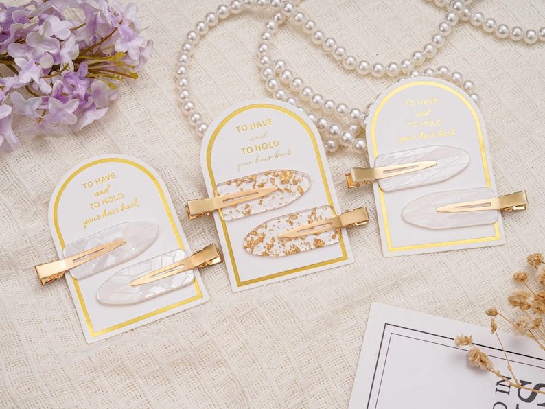 May include: Three sets of hair clips on display. Each set includes two oval-shaped clips with gold-coloured metal accents. The clips are white with gold leaf or a marbled design. The cards read "TO HAVE and TO HOLD your hair back."