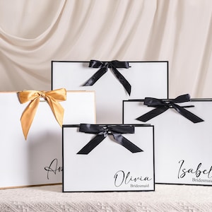 Peut inclure: Plusieurs sacs cadeaux blancs avec des bordures et des nœuds noirs. Un sac a un nœud doré, les autres ont des nœuds noirs. Chaque sac porte un nom en écriture cursive, avec le mot "Bridesmaid" en dessous.