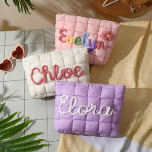 May include: Three quilted cosmetic bags in pink, white, and lilac. Each bag has a name embroidered on it: Evelyn, Chloe, and Elora. The Evelyn bag has a small flower. Heart-shaped sunglasses and a straw hat are in the background.