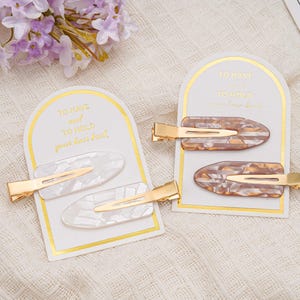 May include: Two sets of hair clips on display. One set features white clips with gold accents, the other has brown and tan clips with gold accents. Each set is presented on a card with gold text that reads "TO HAVE and TO HOLD your hair back."