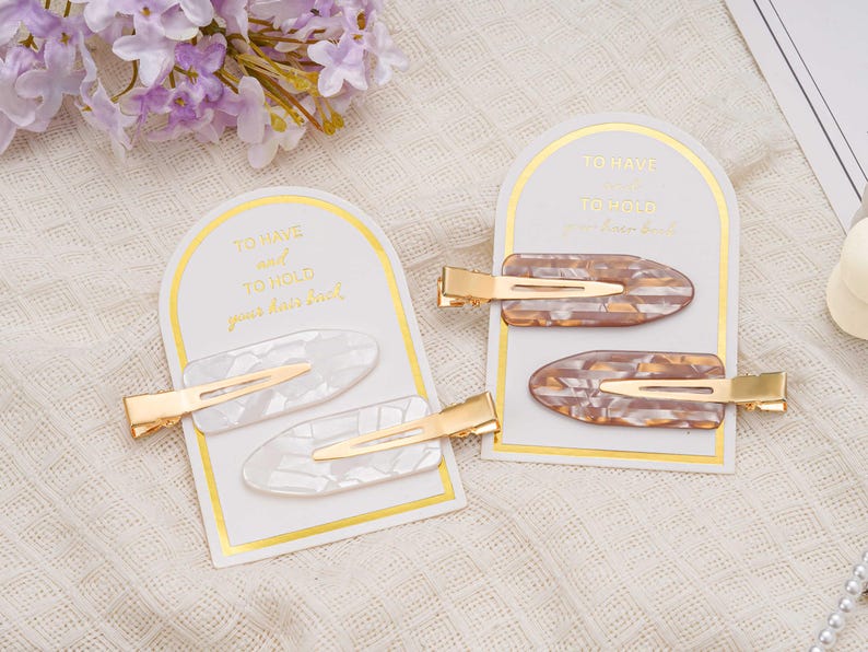 May include: Two sets of hair clips on display. One set features white clips with gold accents, the other has brown and tan clips with gold accents. Each set is presented on a card with gold text that reads "TO HAVE and TO HOLD your hair back."