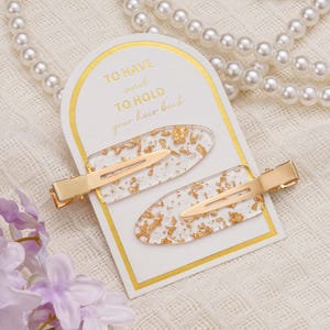 May include: Two gold-coloured hair clips with clear, oval-shaped accents filled with gold leaf flakes. The clips are displayed on a white card with gold trim and the text "TO HAVE and TO HOLD your hair back."