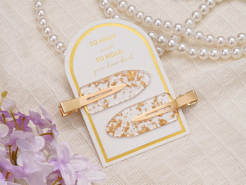 May include: Two gold-coloured hair clips with clear, oval-shaped accents filled with gold leaf flakes. The clips are displayed on a white card with gold trim and the text "TO HAVE and TO HOLD your hair back."
