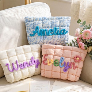 May include: Three quilted pouches, each with a name in yarn. One is blue floral with "Amelia", another cream with "Wendy", and a pink one with "Evelyn". The pouches are on a sofa, with a bouquet of pink flowers in the background.
