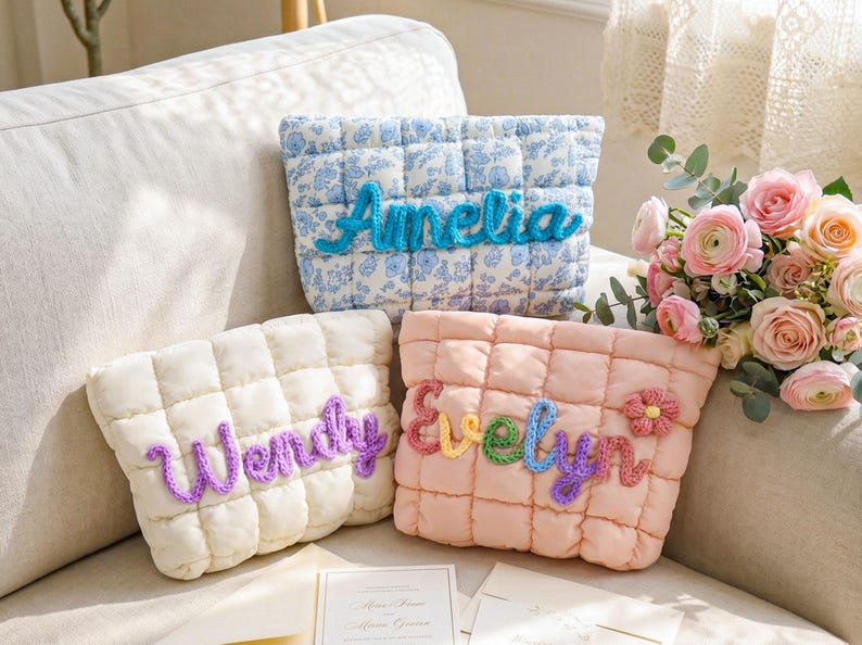 May include: Three quilted pouches, each with a name in yarn. One is blue floral with "Amelia", another cream with "Wendy", and a pink one with "Evelyn". The pouches are on a sofa, with a bouquet of pink flowers in the background.