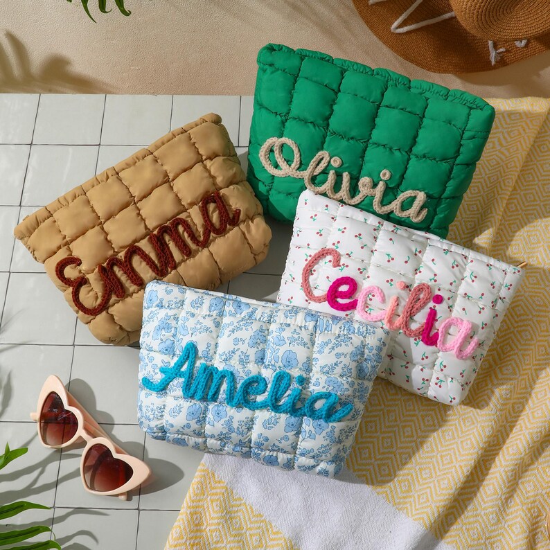 May include: Four quilted cosmetic bags in different colours and patterns, each embroidered with a name: Emma, Olivia, Cecilia, and Amelia. A pair of heart-shaped sunglasses is also visible. The bags are arranged on a patterned surface.