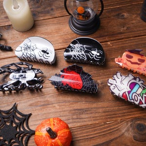 May include: A collection of Halloween-themed hair clips and accessories. Includes spiderweb, spider, and pumpkin designs in black, white, and orange. A decorative candle and lantern are also visible.