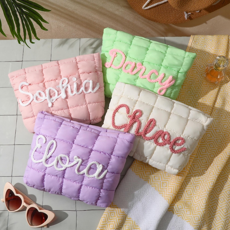 May include: A selection of quilted cosmetic bags in pastel shades: pink, green, cream, and lilac. Each bag has a name embroidered in a contrasting colour: Sophia, Darcy, Chloe, and Elora. The bags are displayed on a beach towel with sunglasses.