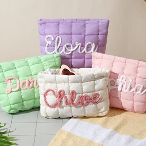 May include: A selection of quilted cosmetic bags in pastel shades: lilac, green, pink, and cream. Each bag has a name embroidered in a contrasting colour: Elora, Dar, Sophia, and Chloe. The bags are displayed on a light-coloured surface.