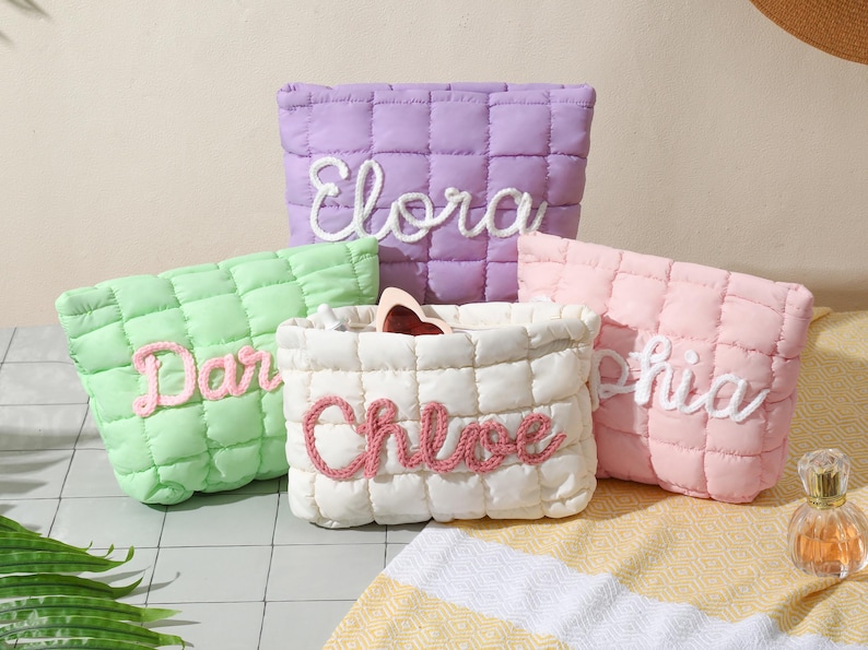 May include: A selection of quilted cosmetic bags in pastel shades: lilac, green, pink, and cream. Each bag has a name embroidered in a contrasting colour: Elora, Dar, Sophia, and Chloe. The bags are displayed on a light-coloured surface.