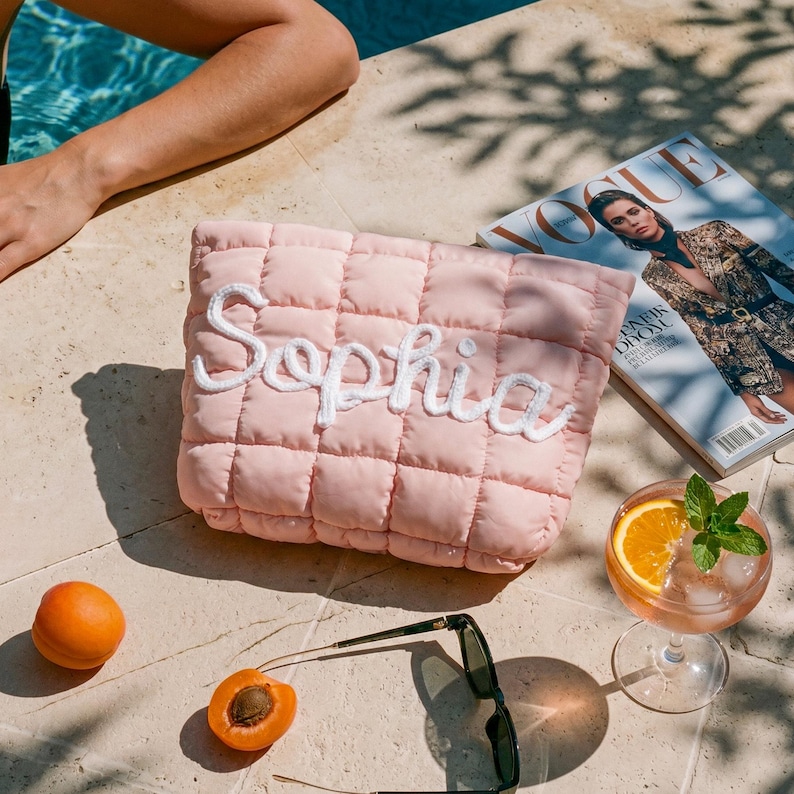 May include: A pink quilted clutch bag embroidered with the name "Sophia" in white. The bag is rectangular and rests on a stone surface. Nearby are a Vogue magazine, sunglasses, a cocktail, and apricots.
