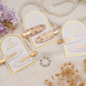 May include: Oval hair clips in tortoiseshell and white, with gold-coloured metal clasps. Each pair is displayed on a white card with gold trim and the text "TO HAVE and TO HOLD your hair back".