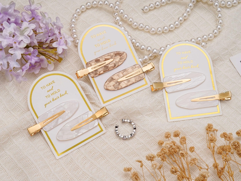 May include: Oval hair clips in tortoiseshell and white, with gold-coloured metal clasps. Each pair is displayed on a white card with gold trim and the text "TO HAVE and TO HOLD your hair back".