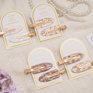 May include: Four sets of hair clips are displayed. Each set contains two decorative clips with gold-coloured metal clasps. The clips feature oval designs in white, gold, and brown tones, presented on cards with the text "TO HAVE and TO HOLD your hair back."
