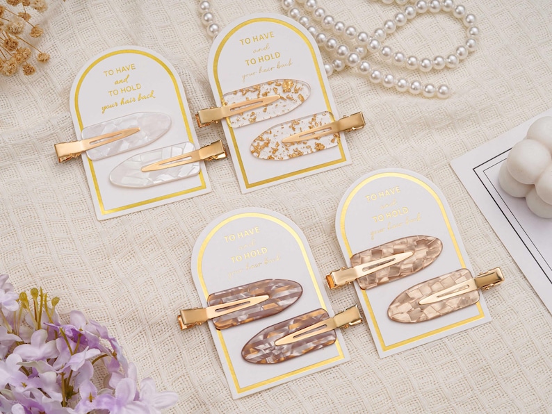 May include: Four sets of hair clips are displayed. Each set contains two decorative clips with gold-coloured metal clasps. The clips feature oval designs in white, gold, and brown tones, presented on cards with the text "TO HAVE and TO HOLD your hair back."