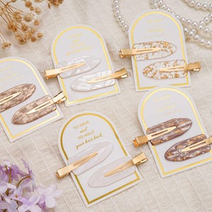 May include: Multiple sets of hair clips are presented. Each set features two oval clips with gold-coloured metal clasps. The clips display a marbled or speckled pattern in shades of white, brown, and gold. The cards read "TO HAVE and TO HOLD your hair back."