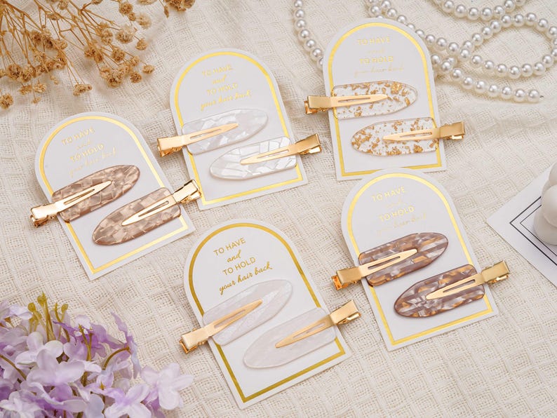 May include: Multiple sets of hair clips are presented. Each set features two oval clips with gold-coloured metal clasps. The clips display a marbled or speckled pattern in shades of white, brown, and gold. The cards read "TO HAVE and TO HOLD your hair back."