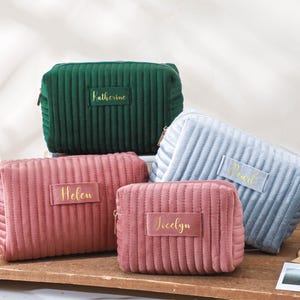 Puede incluir: Varias bolsas de cosméticos personalizadas en tonos verdes, rosas y azules. Cada bolsa tiene un diseño acanalado y una etiqueta dorada con un nombre. Las bolsas están dispuestas sobre una superficie de madera.