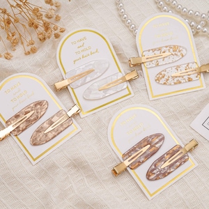 May include: Four sets of hair clips are presented. Each set contains two oval clips with gold-coloured metal clasps. The clips display patterns in white, gold, and brown marbled designs. The cards read "TO HAVE and TO HOLD your hair back."