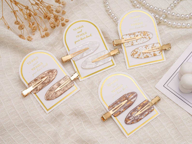 May include: Four sets of hair clips are presented. Each set contains two oval clips with gold-coloured metal clasps. The clips display patterns in white, gold, and brown marbled designs. The cards read "TO HAVE and TO HOLD your hair back."