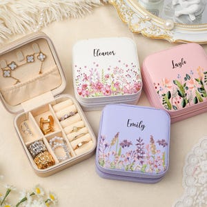 Può includere: Un portagioie beige aperto con anelli, orecchini e bracciali. Tre portagioie quadrati personalizzati in bianco, rosa e lavanda, ciascuno con un design floreale e un nome: Eleanor, Layla e Emily.
