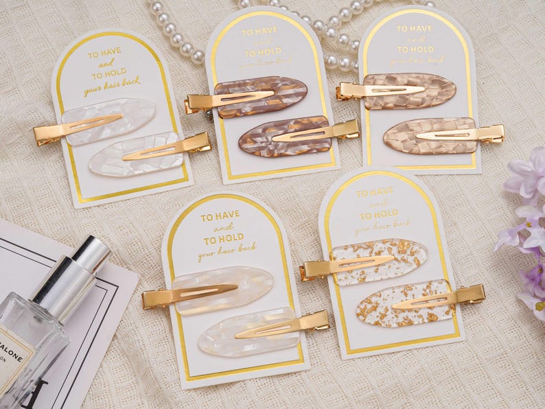 May include: A selection of hair clips with gold-coloured clasps and various patterns, including white, brown, and gold. Each pair of clips is presented on a card with the text "TO HAVE and TO HOLD your hair back."