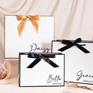Peut inclure: Trois sacs cadeaux blancs avec des bordures noires et des nœuds en satin. Le plus grand sac a un nœud doré, tandis que les deux autres ont des nœuds noirs. Chaque sac porte un nom : Daisy, Bella et Grace.