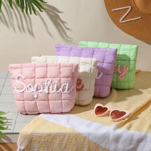 May include: Four quilted cosmetic bags in pastel shades: pink, cream, lilac, and green. Each bag has a name embroidered in a cursive font. Heart-shaped sunglasses sit on a yellow and white striped towel. A straw hat with a white letter "Z" is in the background.