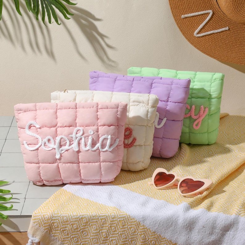 May include: Four quilted cosmetic bags in pastel shades: pink, cream, lilac, and green. Each bag has a name embroidered in a cursive font. Heart-shaped sunglasses sit on a yellow and white striped towel. A straw hat with a white letter "Z" is in the background.
