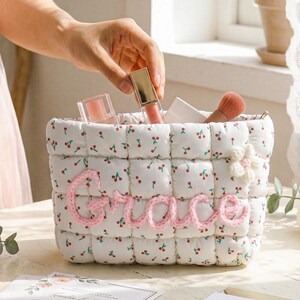 May include: A white quilted cosmetic bag with a cherry pattern and the name "Grace" in pink yarn. The bag contains makeup items, including lip gloss and a blush brush. A hand is reaching into the bag.