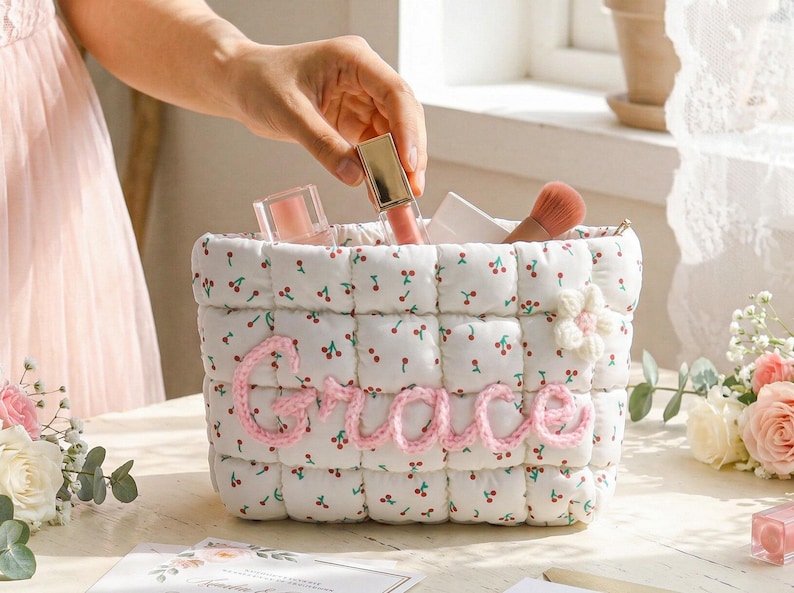 May include: A white quilted cosmetic bag with a cherry pattern and the name "Grace" in pink yarn. The bag contains makeup items, including lip gloss and a blush brush. A hand is reaching into the bag.