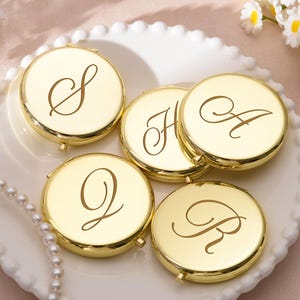 Puede incluir: Espejos compactos dorados con letras grabadas S, H, A, Q y R, dispuestos sobre un plato blanco con borde festoneado. Los espejos son redondos y tienen un acabado brillante. Un collar de perlas y flores en el fondo.