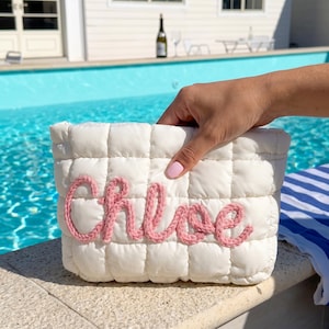 May include: A white, quilted cosmetic bag with the name "Chloe" embroidered in pink yarn. The bag is held near a swimming pool, suggesting a summer theme. The bag has a rectangular shape.