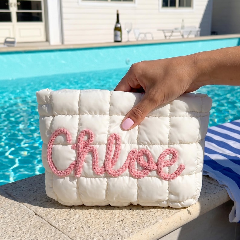 May include: A white, quilted cosmetic bag with the name "Chloe" embroidered in pink yarn. The bag is held near a swimming pool, suggesting a summer theme. The bag has a rectangular shape.