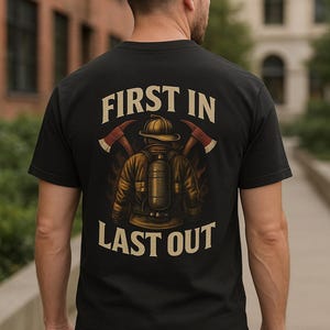 May include: Black t-shirt with a firefighter graphic on the back. The design features a firefighter with axes and the text "FIRST IN LAST OUT" in a bold, vintage font. The shirt is worn by a person in blue jeans.