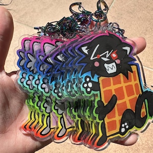 May include: A stack of clear acrylic keychains featuring a cartoon black cat holding a waffle. The cat has red eyes and a wide grin. The keychains have rainbow gradient edges and iridescent metal key rings.
