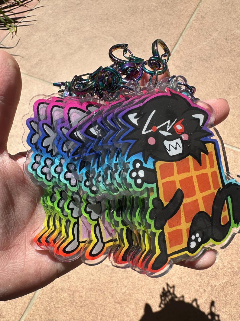 May include: A stack of clear acrylic keychains featuring a cartoon black cat holding a waffle. The cat has red eyes and a wide grin. The keychains have rainbow gradient edges and iridescent metal key rings.