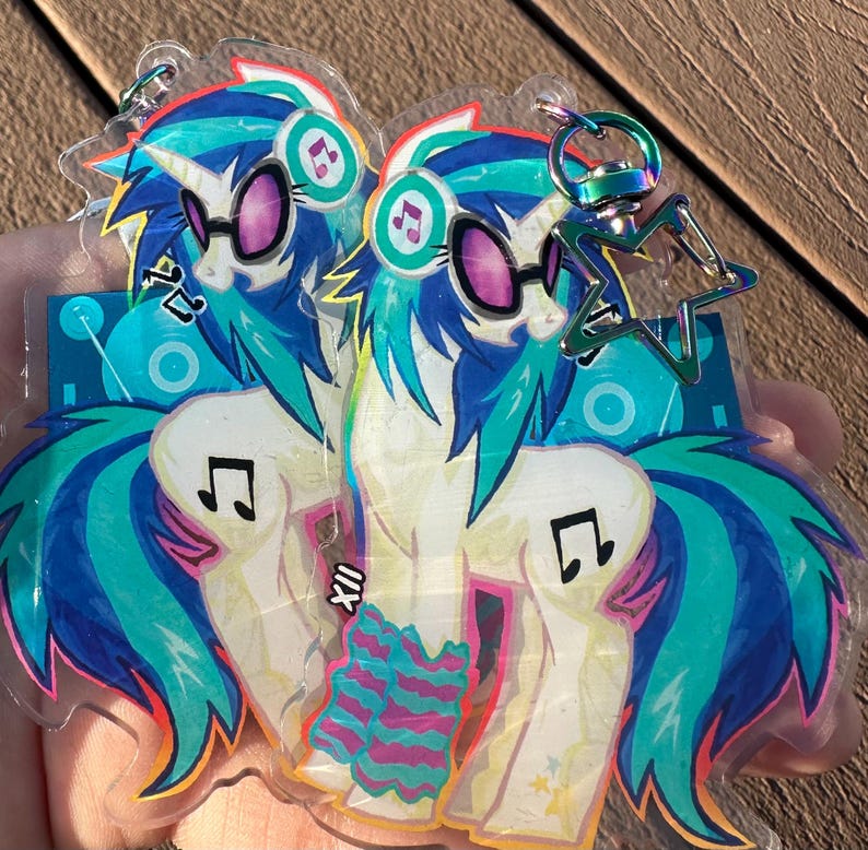 Pode incluir: Um par de chaveiros de acr&iacute;lico transparente com um design de p&ocirc;nei de desenho animado. Os p&ocirc;neis t&ecirc;m cabelo azul e verde-azulado, &oacute;culos de sol e detalhes de notas musicais. Cada chaveiro tem um fecho em forma de estrela.