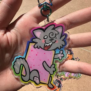 May include: A clear acrylic keychain featuring a cartoon cat holding a pink rectangular pastry. The cat is gray with a smiling face and a rainbow outline. The keychain has a star-shaped iridescent clasp.