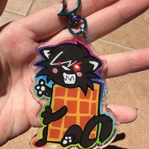 May include: A clear acrylic keychain featuring a cartoon black cat with red eyes and a waffle. The cat has white teeth and is outlined in black with a rainbow border. The keychain has a silver clasp.