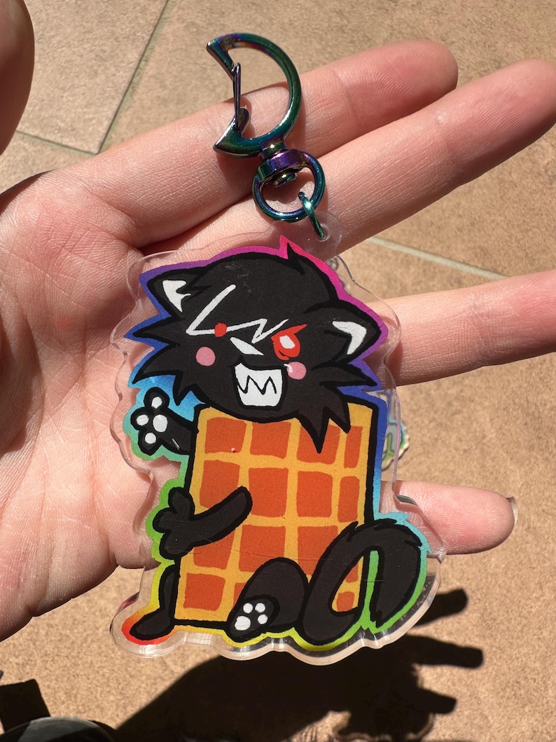May include: A clear acrylic keychain featuring a cartoon black cat with red eyes and a waffle. The cat has white teeth and is outlined in black with a rainbow border. The keychain has a silver clasp.