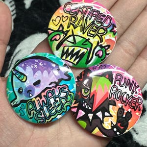 May include: Three colorful round buttons with unique designs. One says "Certified Raver" with a monster graphic, another says "Always Sleep" with a narwhal, and the last says "Punk Rocker" with a monster graphic.