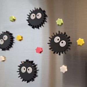 May include: A collection of black, spiky magnets with large white eyes, resembling soot sprites, are scattered across a metallic surface. Colorful star-shaped magnets in yellow, pink, green, and white are also present, creating a playful design.