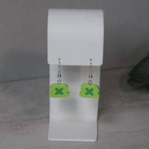 May include: A pair of lime green earrings with a dark green X design. The earrings are displayed on a white stand. The earrings have silver-colored hooks.