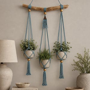 May include: Three hanging macrame planters with faux greenery. The planters are suspended from a natural wood branch with blue macrame rope and gold accents. Each planter has a tassel. The planters are on a wooden table with a lamp and decorative items.